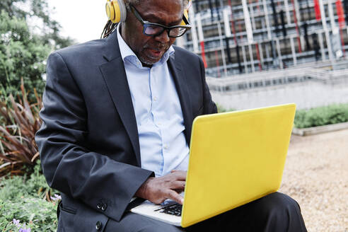Male professional with headphones using laptop while sitting at garden - ASGF00928