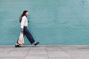 Junge Frau mit Netztasche auf Fußweg an türkisfarbener Backsteinmauer - ASGF00953