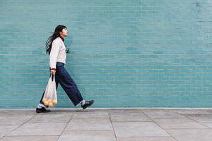 Young woman with mesh bag on footpath by turquoise brick wall - ASGF00953