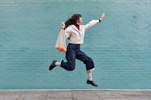 Excited young woman jumping while holding mesh bag by turquoise brick wall - ASGF00955