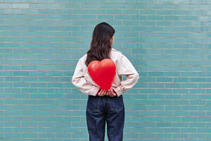 Woman holding heart shape balloon behind her back in front of turquoise brick wall - ASGF00965