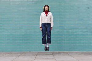 Woman with headphones levitating in front of turquoise brick wall - ASGF00967