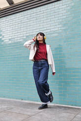 Woman listening music while standing on one leg in front of turquoise brick wall - ASGF00971