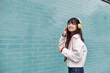 Smiling young beautiful woman with headphones standing by brick wall - ASGF00976