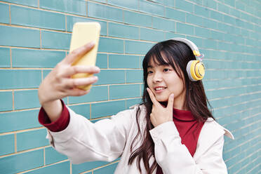 Woman taking selfie through smart phone by brick wall - ASGF00978