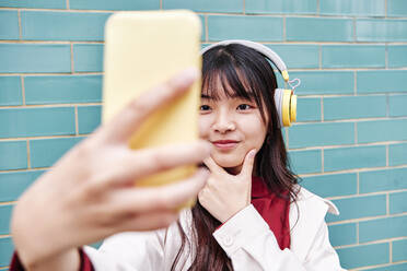 Woman with headphones taking selfie in front of brick wall - ASGF00979