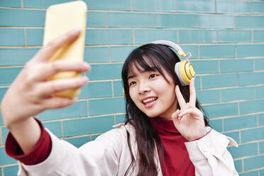 Woman showing peace sign while taking selfie in front of turquoise brick wall - ASGF00980