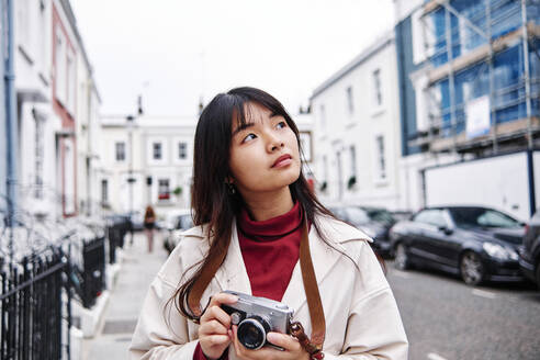 Thoughtful woman holding camera in city - ASGF00983