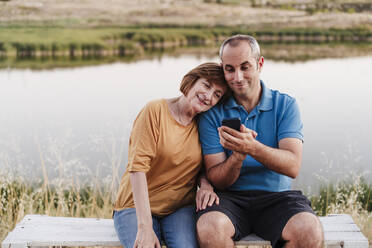 Sohn benutzt sein Smartphone, während er mit seiner Mutter auf einer Bank am See sitzt - EBBF04408