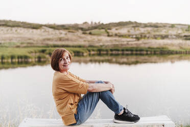 Senior woman sitting on bench by lake - EBBF04411