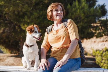 Lächelnde ältere Frau schaut weg, während sie mit ihrem Hund auf einer Bank sitzt - EBBF04415