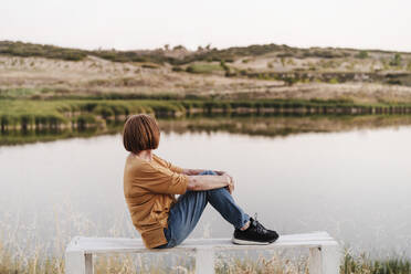 Senior woman looking at mountains while sitting on bench near lake - EBBF04427