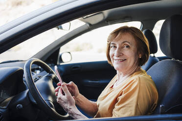 Lächelnde ältere Frau hält ihr Smartphone im Auto sitzend - EBBF04436