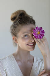 Smiling young woman covering eye with purple flower in studio - EIF01807