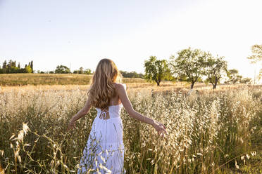 Blonde junge Frau in weißem Kleid geht inmitten von Gras auf einem Feld - EIF01860