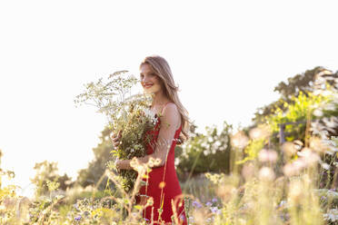 Smiling blond woman looking away while holding wildflowers bouquet in field - EIF01864