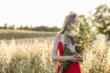Junge blonde Frau riecht an einem Strauß Wildblumen auf einem Feld - EIF01879