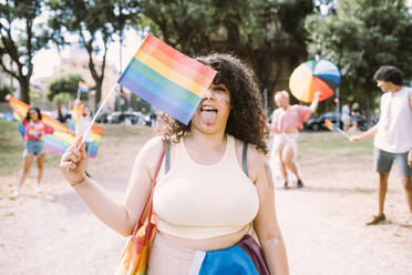 Junge Frau hält Regenbogenflagge, während Freunde im Hintergrund im Park - MEUF03621