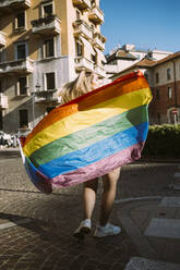Young female activist with rainbow flag walking on street during sunny day - MEUF03686