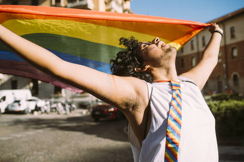 Young male activist looking up while holding rainbow flag on street - MEUF03689