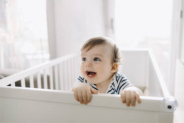 Cute baby boy looking away in crib at home - GMCF00231