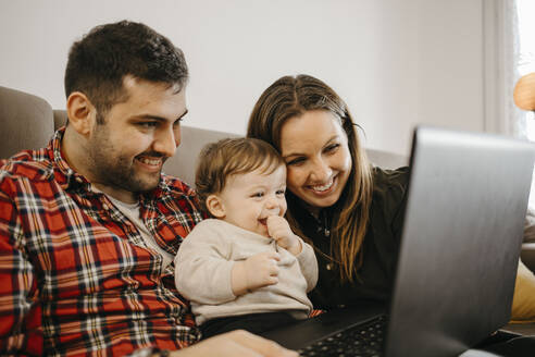 Glückliche Mutter und Vater mit Sohn während eines Videogesprächs auf dem Laptop im Wohnzimmer sitzend - GMCF00236