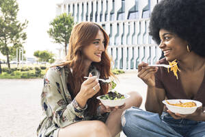 Smiling young women having food while talking during sunny day - JCCMF03376