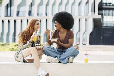 Female friends enjoying food while sitting on bench - JCCMF03384