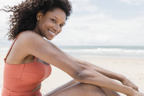 Glückliche junge Frau im Badeanzug am Strand sitzend - JRVF01269