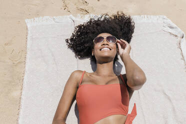 Lächelnde junge Frau mit lockigem Haar und Sonnenbrille, die auf einem Strandtuch liegt - JRVF01275