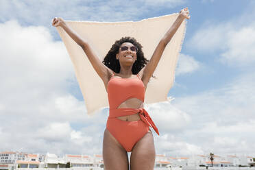 Lächelnde Frau mit Sonnenbrille hält Strandtuch an einem sonnigen Tag - JRVF01280