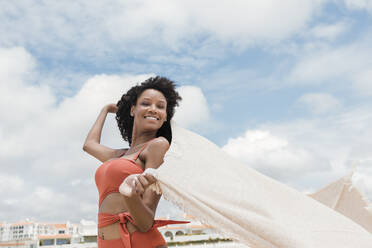 Lächelnde schöne junge Frau hält Strandtuch an einem sonnigen Tag - JRVF01283