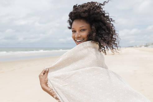 Smiling young woman with towel looking over shoulder at beach - JRVF01285