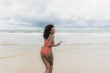 Cheerful woman running with mobile phone at beach - JRVF01302