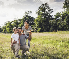 Father holding airplane toy while crouching by son and woman on grass - UUF24214