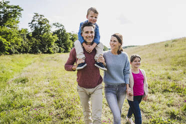 Father carrying son on shoulders while walking with family at meadow - UUF24243