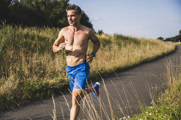 Shirtless sportsman running on road during sunny day - UUF24250