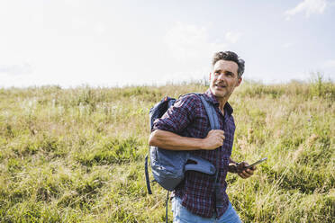 Männlicher Wanderer mit Rucksack und Mobiltelefon auf einer Wiese - UUF24294