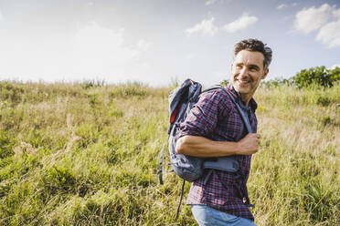 Lächelnder Mann mit Rucksack beim Wandern auf einer Wiese an einem sonnigen Tag - UUF24295