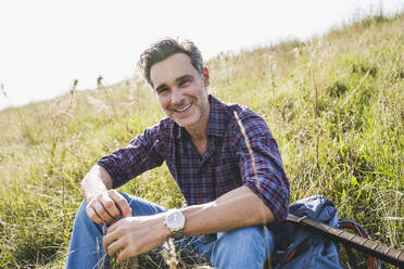 Smiling man sitting with backpack and guitar on grass - UUF24297