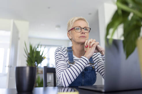 Thoughtful businesswoman with hands clasped sitting at desk in home office - WPEF05127