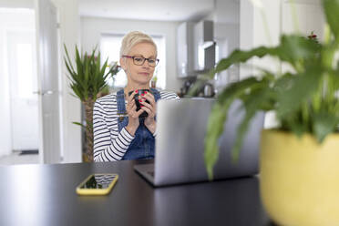 Reife Frau, die eine Tasse hält, während sie im Heimbüro auf einen Laptop schaut - WPEF05128