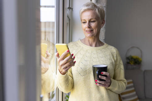 Reife Frau, die ein Smartphone benutzt und eine Tasse zu Hause hält - WPEF05135