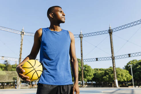 Mann schaut weg, während er einen Basketball auf einem Sportplatz an einem sonnigen Tag hält - XLGF02120