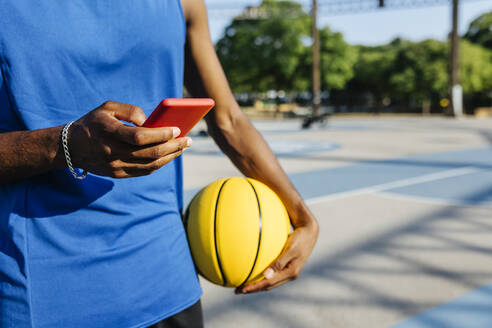 Mann, der ein Smartphone benutzt, während er einen Basketball auf einem Sportplatz hält - XLGF02133