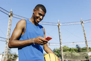 Smiling young man using smart phone at basketball court - XLGF02134