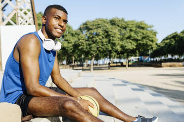 Male basketball player with headphones looking away while sitting on steps at sports court - XLGF02136