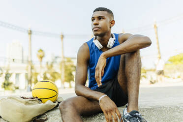 Young male basketball player looking away while sitting at sports court - XLGF02137