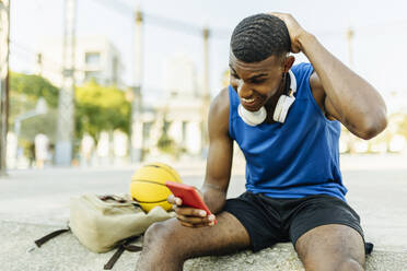 Young man with hand behind head smiling while using smart phone at basketball court - XLGF02138