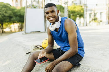 Smiling man holding smart phone while sitting at basketball court - XLGF02139